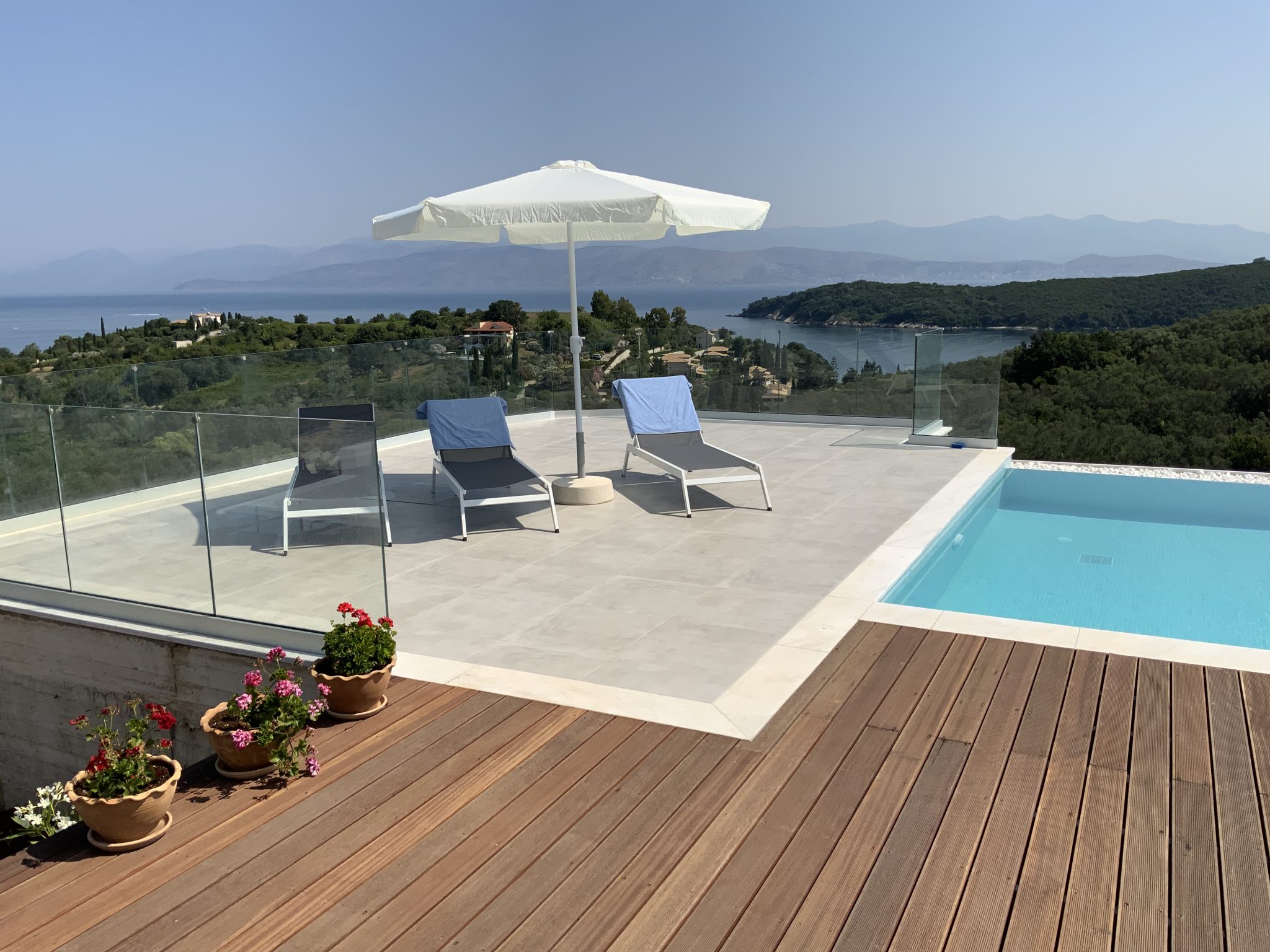 A rooftop pool area with two lounge chairs under an umbrella, overlooking a coastal landscape with mountains and greenery in the distance. Potted plants adorn the wooden deck, completing this idyllic scene of villa rentals in Corfu.