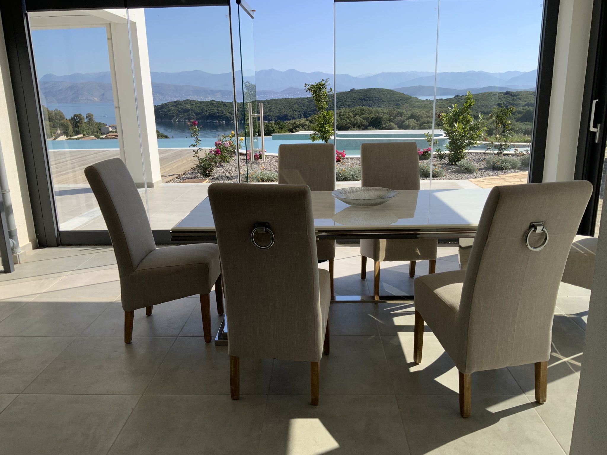 A dining area with a glass table and six beige upholstered chairs, reminiscent of Corfu luxury villas, set against large windows with a view of a pool, trees, hills, and distant mountains.