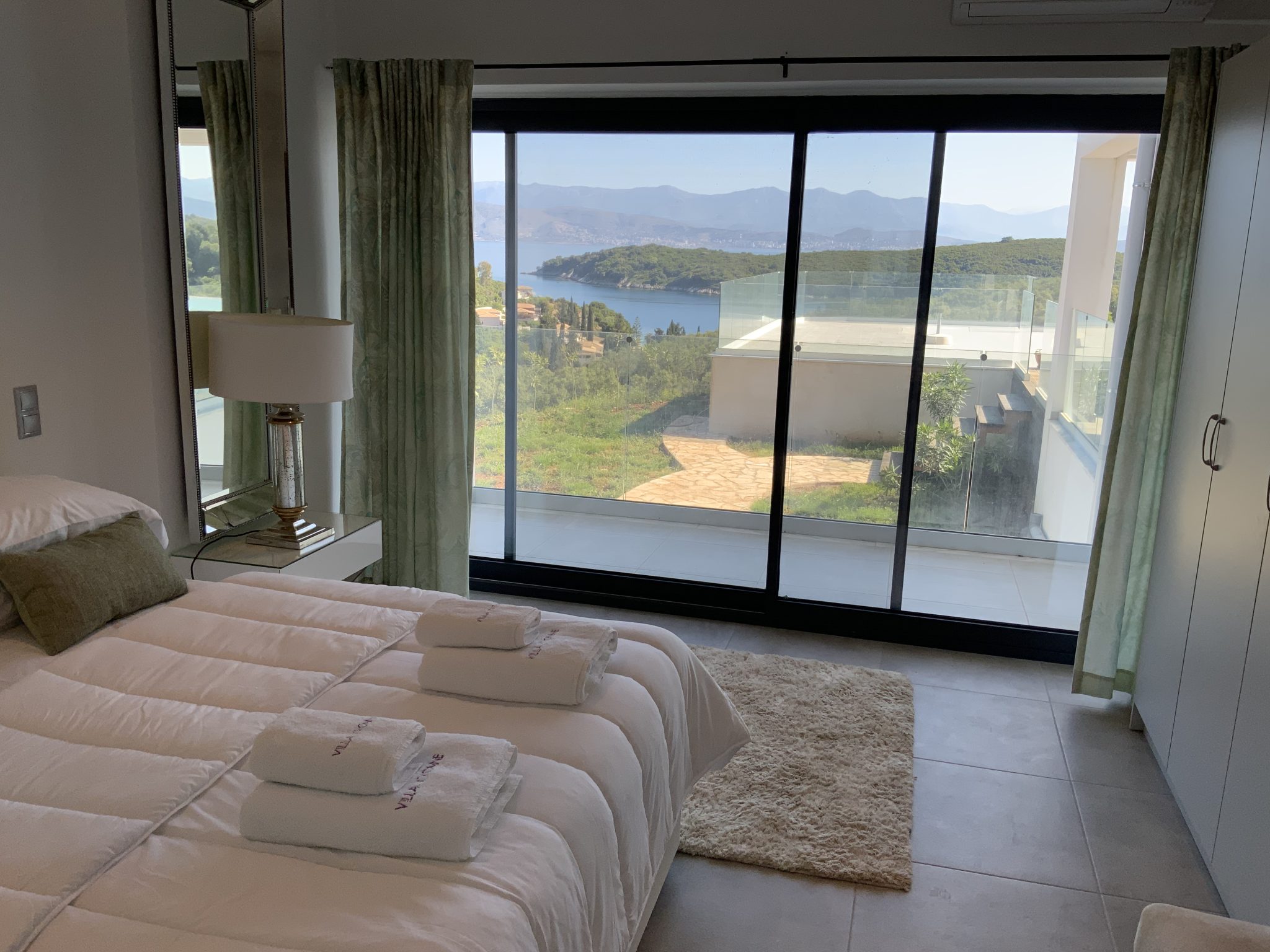 A bedroom in one of the luxury villas in Corfu, featuring a made bed with two folded towels on top, a small side table with a lamp, and a large window offering a scenic view of mountains and water.