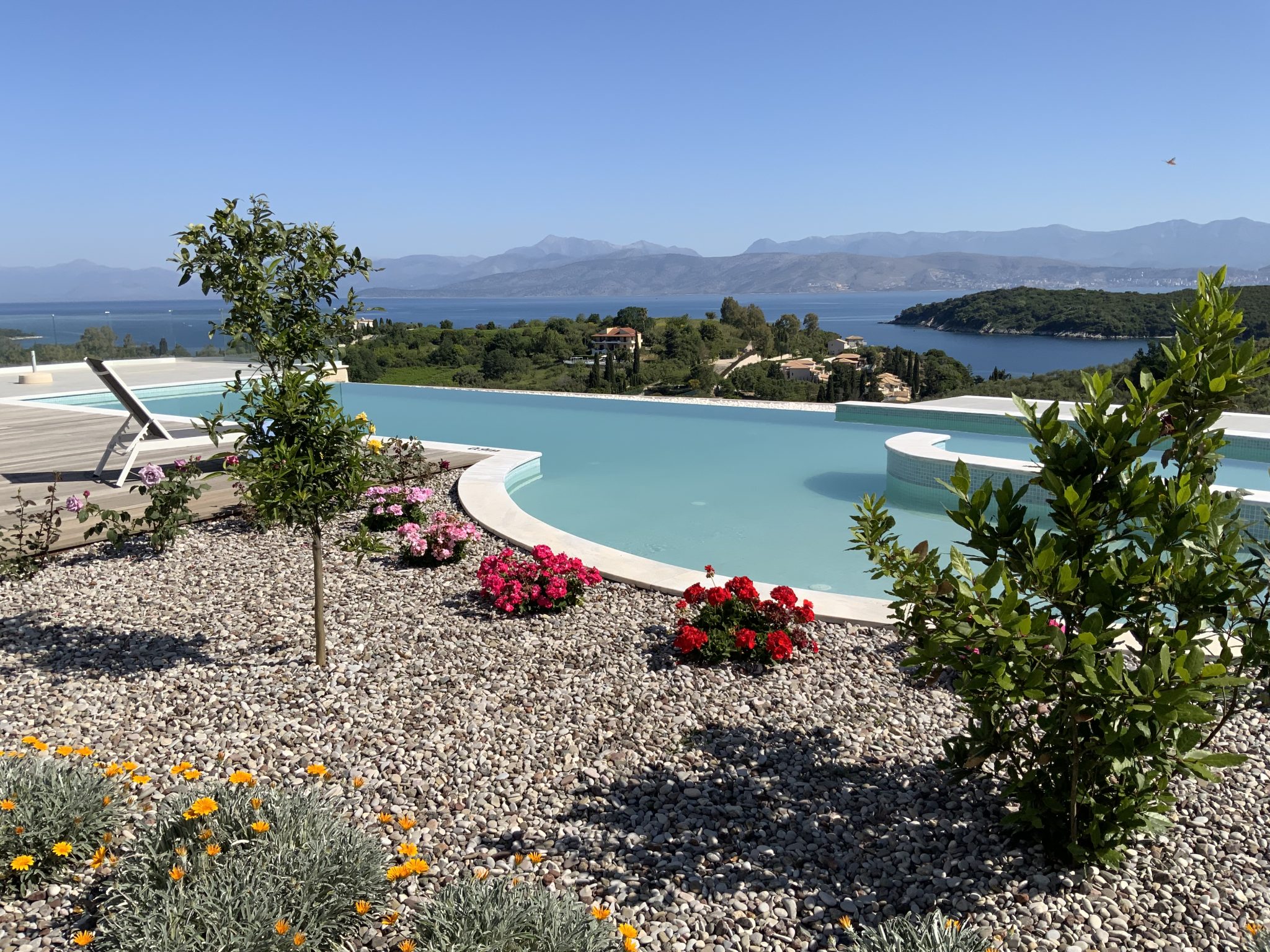 Infinity pool surrounded by lush plants and vibrant flowers, with a view of towering mountains and sparkling water in the background—experience this serene escape at one of the beautiful Corfu villas to rent.