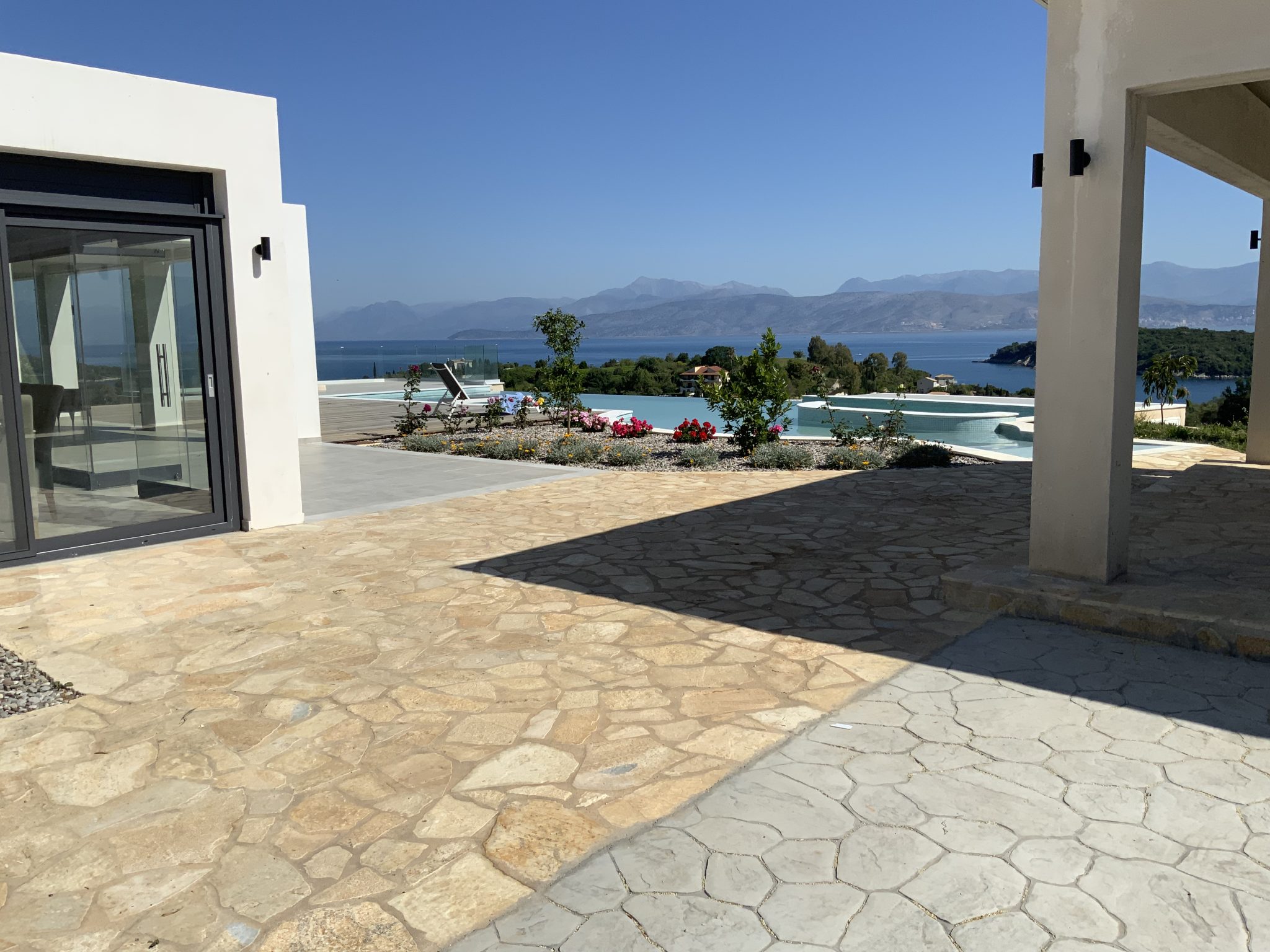 A stone patio in one of the luxury Corfu villas overlooks a swimming pool and the ocean, with a clear blue sky overhead and mountains visible in the distance.