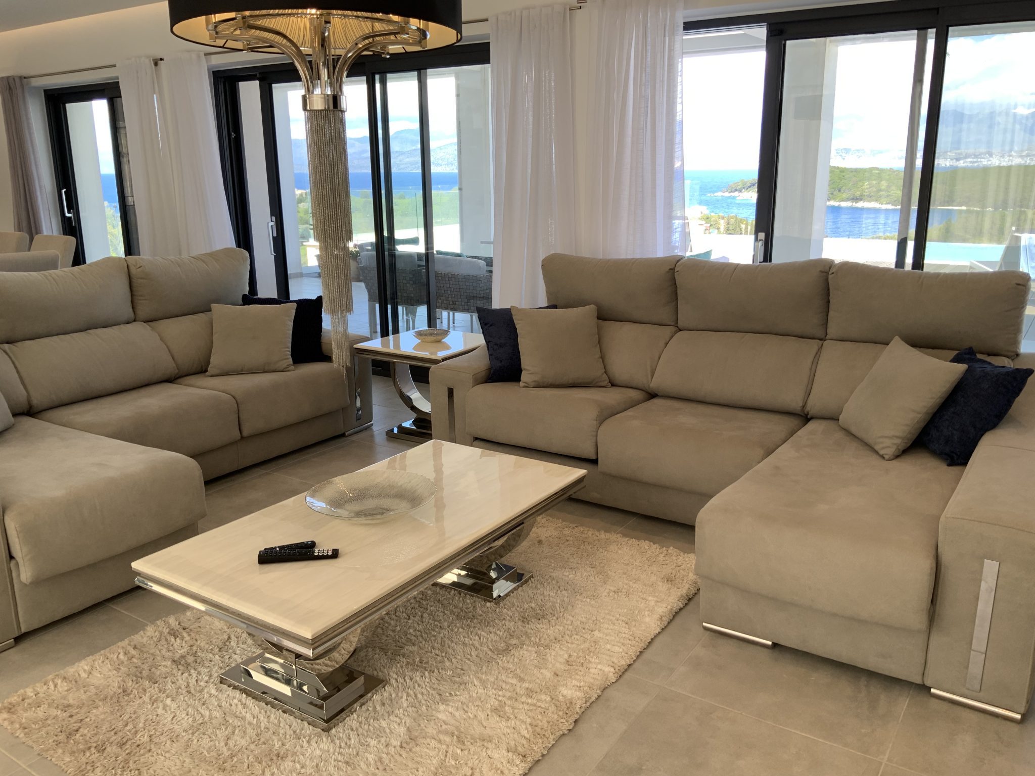 A modern living room in a luxury villa in Corfu with two beige sofas, a coffee table, and a wall-mounted flat-screen TV. Large windows provide a view of a scenic landscape, and white curtains are partially drawn.