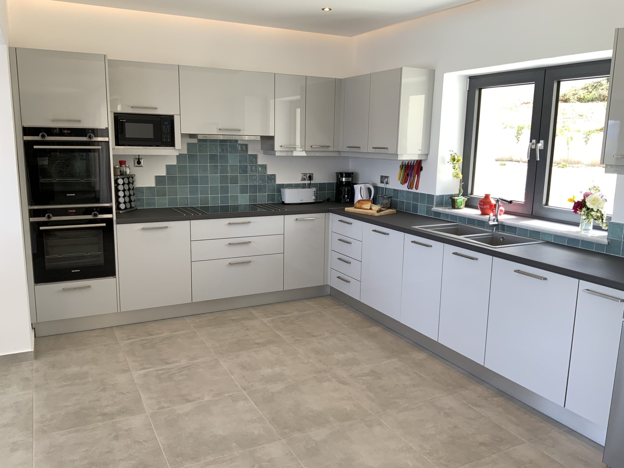 Modern kitchen with white cabinets, black countertop, and appliances including a double oven, microwave, and coffee maker. Features a tiled backsplash and a window above the sink offering serene views reminiscent of a Corfu villa holiday.