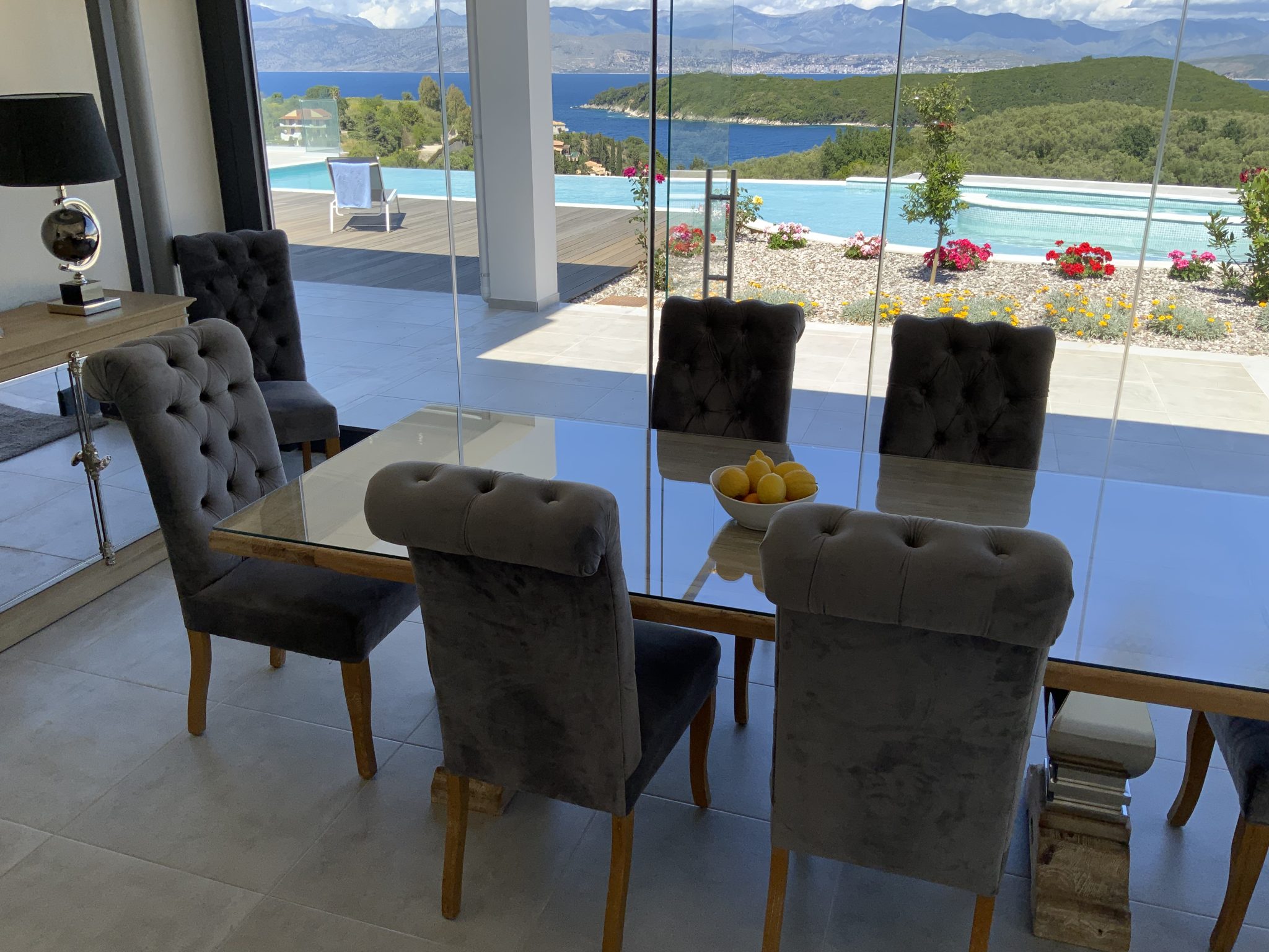 Glass dining table with six gray upholstered chairs in a modern room, overlooking a balcony with a pool and scenic view of a mountainous landscape and a lake. A bowl of lemons sits on the table, offering a taste of luxury akin to villa rentals in Corfu.