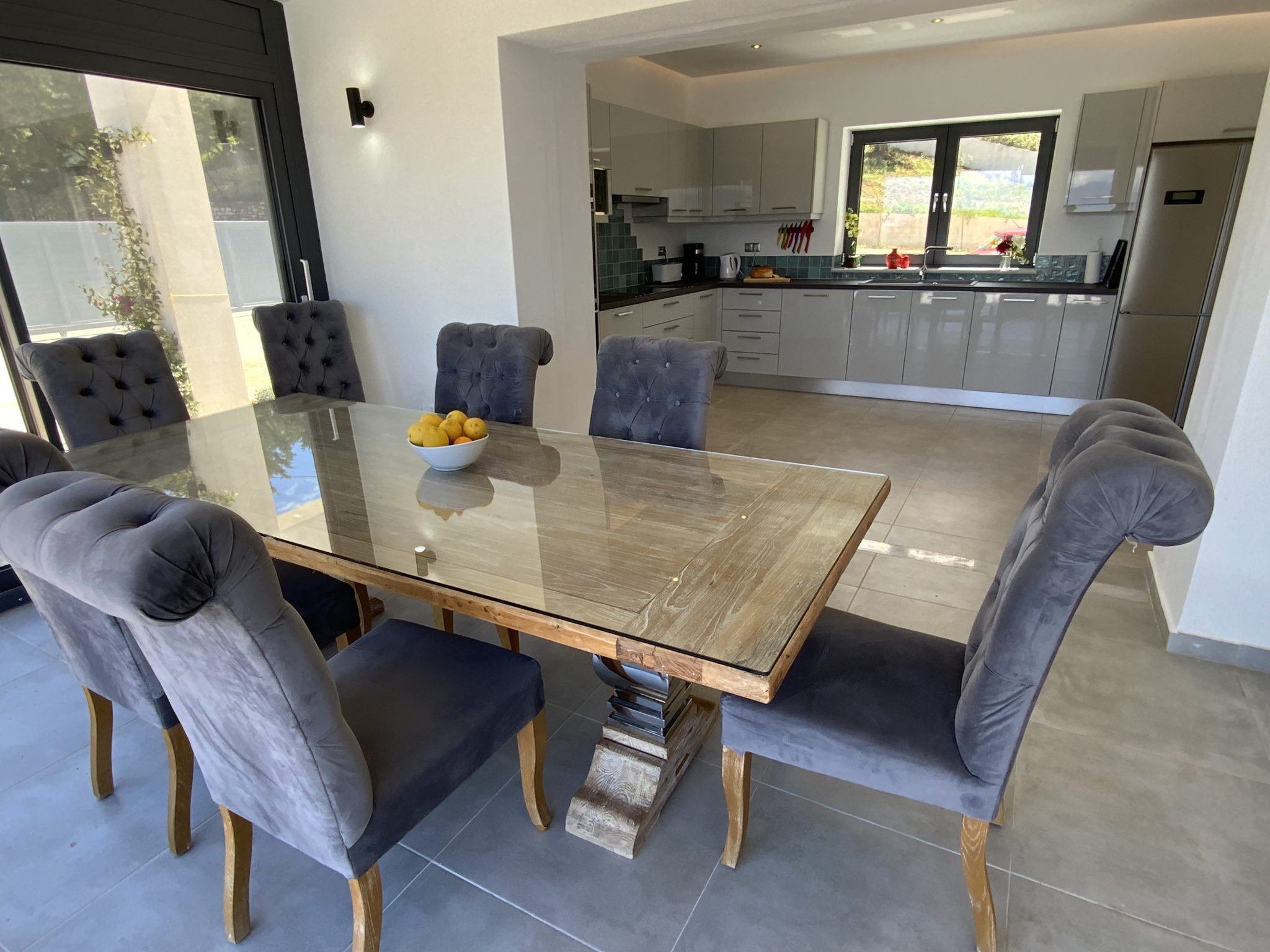 A modern dining area with a glass-topped wooden table and six gray upholstered chairs in a luxury villa rental in Greece. A bowl of lemons sits on the table, and the background features an open kitchen with gray cabinets and appliances.