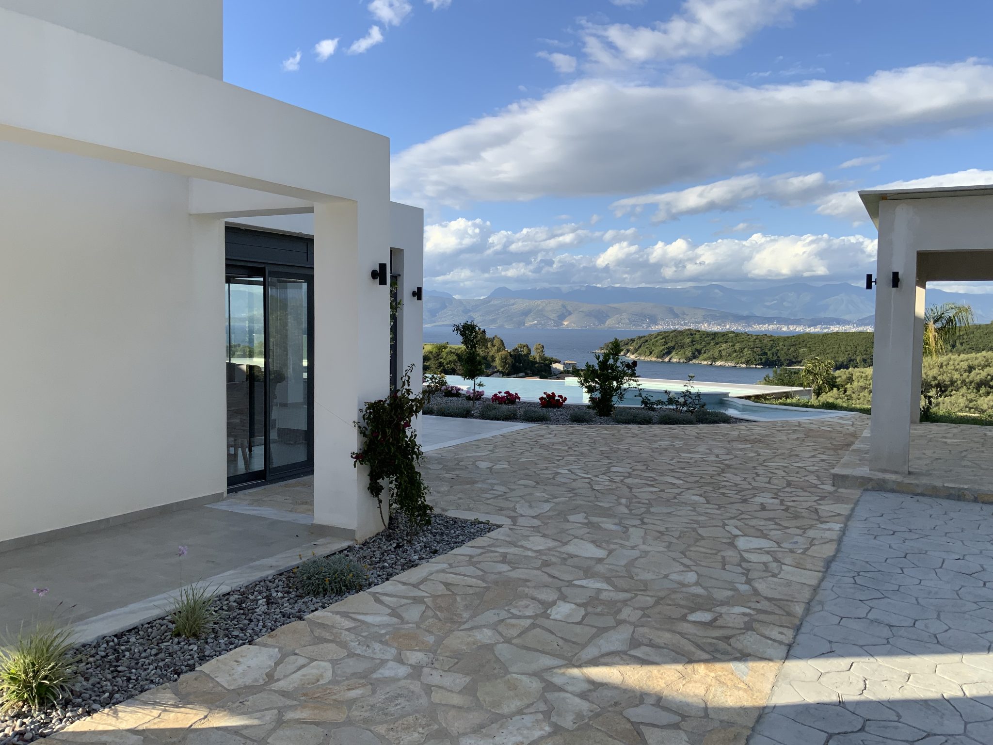 Modern building with white walls and a stone patio overlooking a scenic landscape with mountains, greenery, and a body of water under a partly cloudy sky. Perfect for those seeking villa rentals in Corfu.