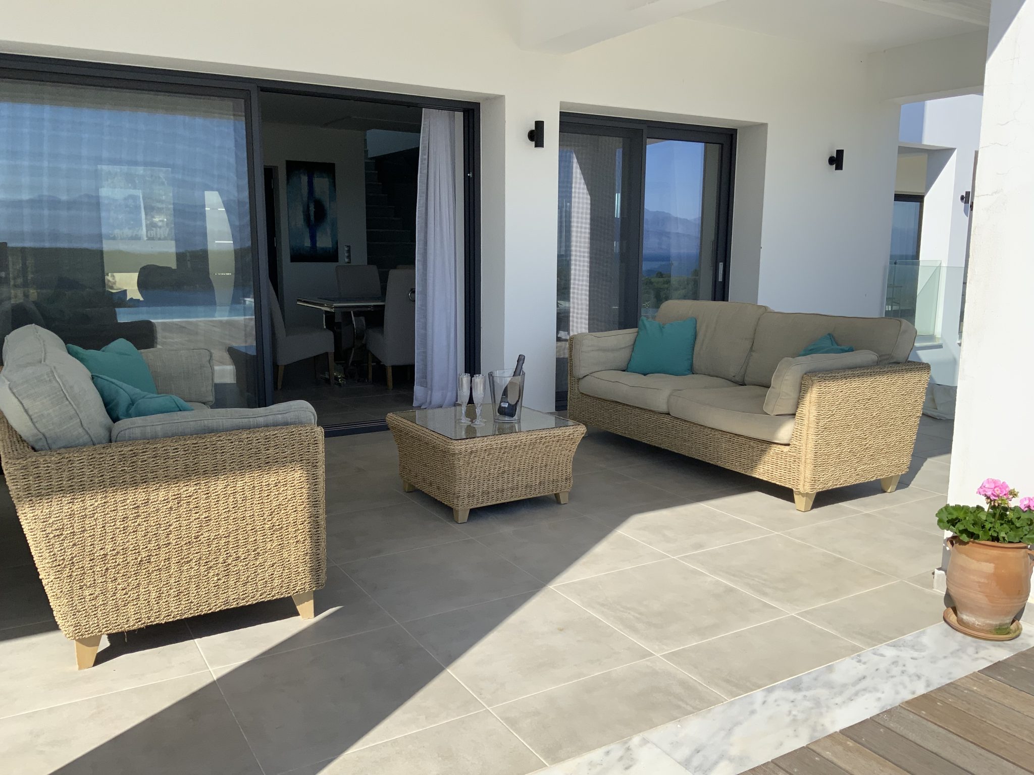 Patio with wicker furniture, including two sofas and a glass-top table, adjacent to a sliding glass door. A potted plant sits on the right, and mountains are visible through the window. This serene spot is reminiscent of elegant villas in Corfu.