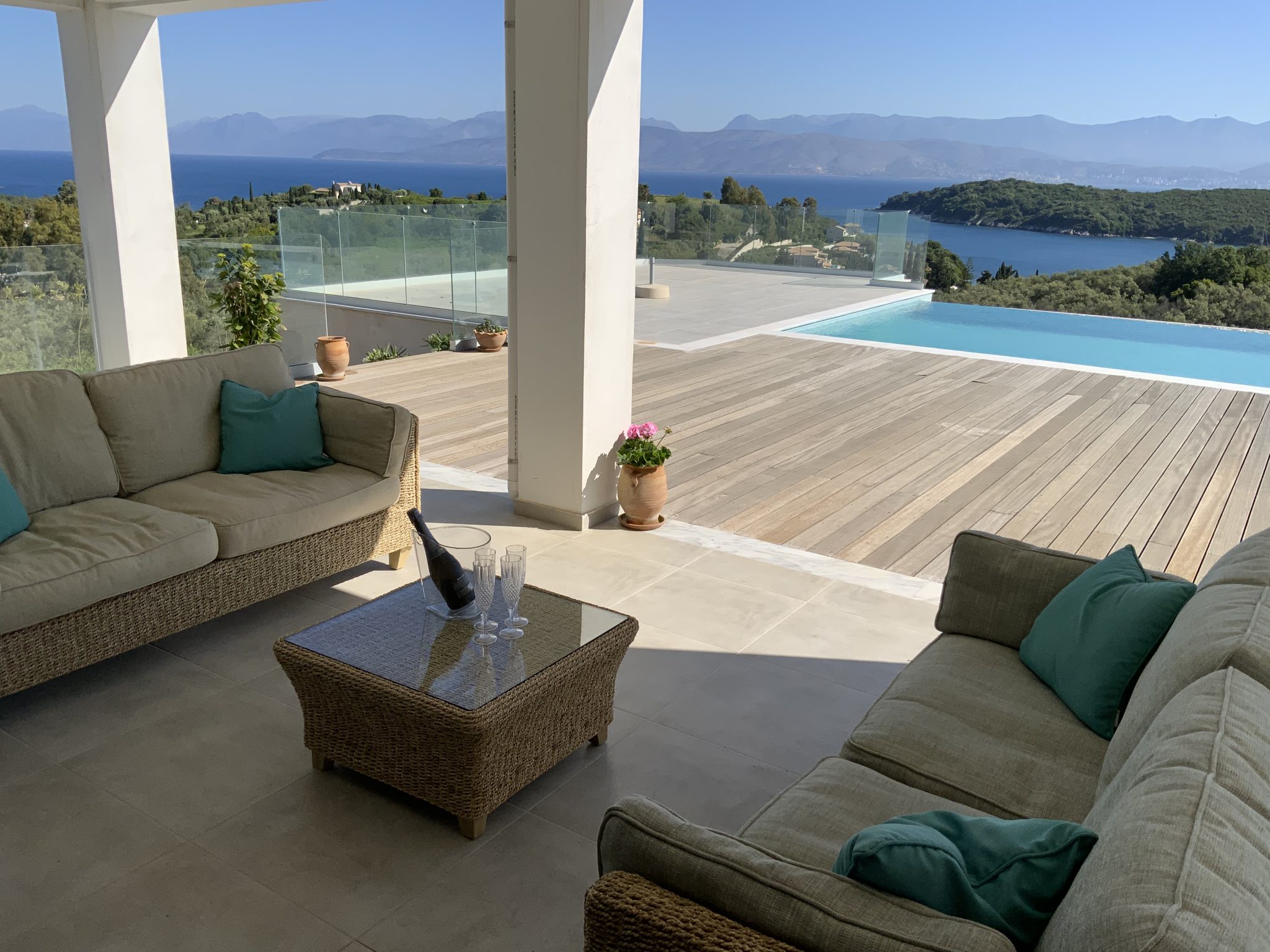 A patio with two beige wicker sofas, a glass coffee table, and views of an infinity pool, lush greenery, and the clear blue ocean in the background - a perfect example of villas in Corfu with private pools.