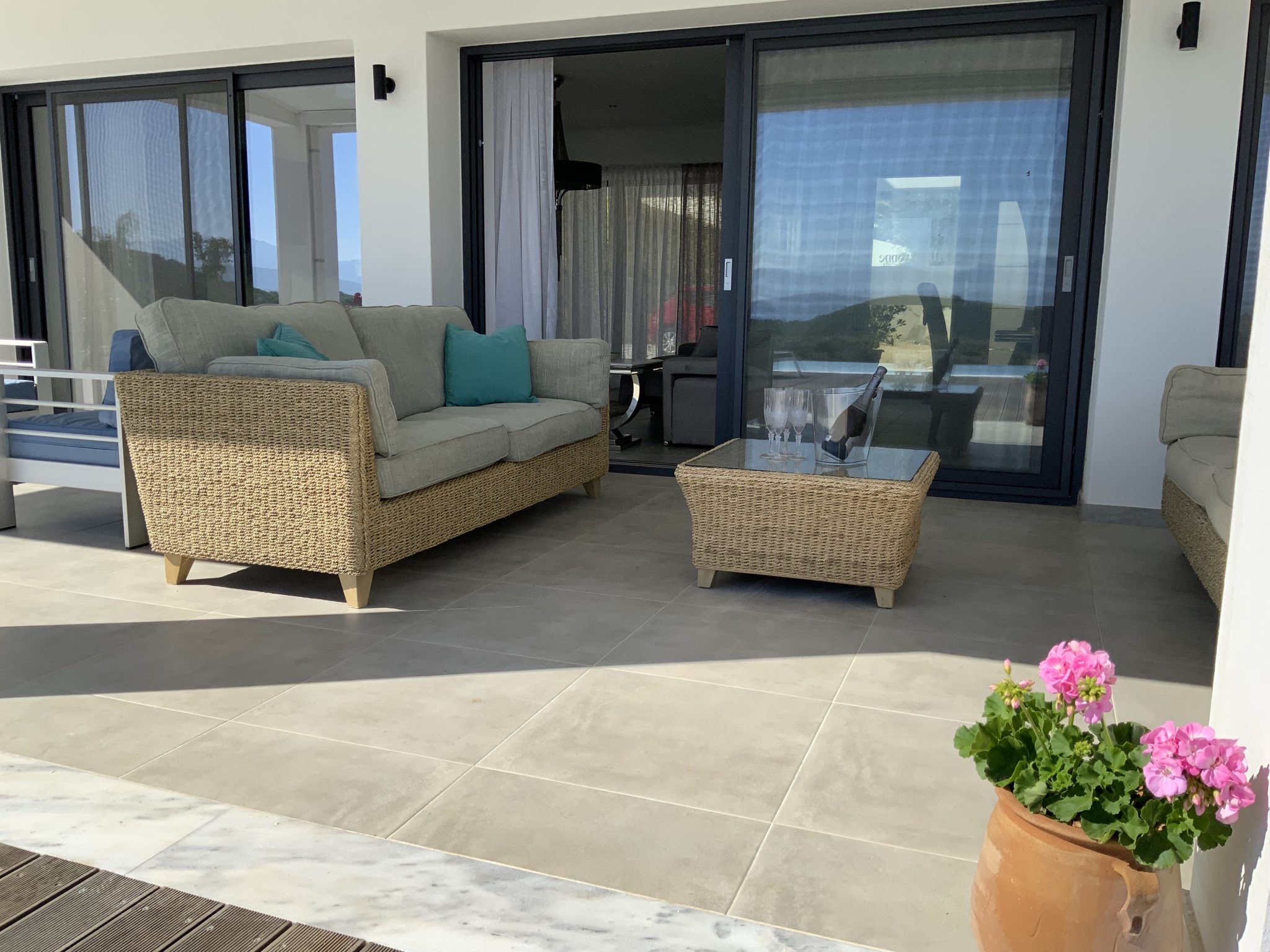 A patio with a gray tile floor features wicker furniture including a sofa, chairs, and a coffee table. Glass sliding doors open to an interior. A pink flower in a pot is in the foreground, evoking the charm of villa rentals in Corfu.