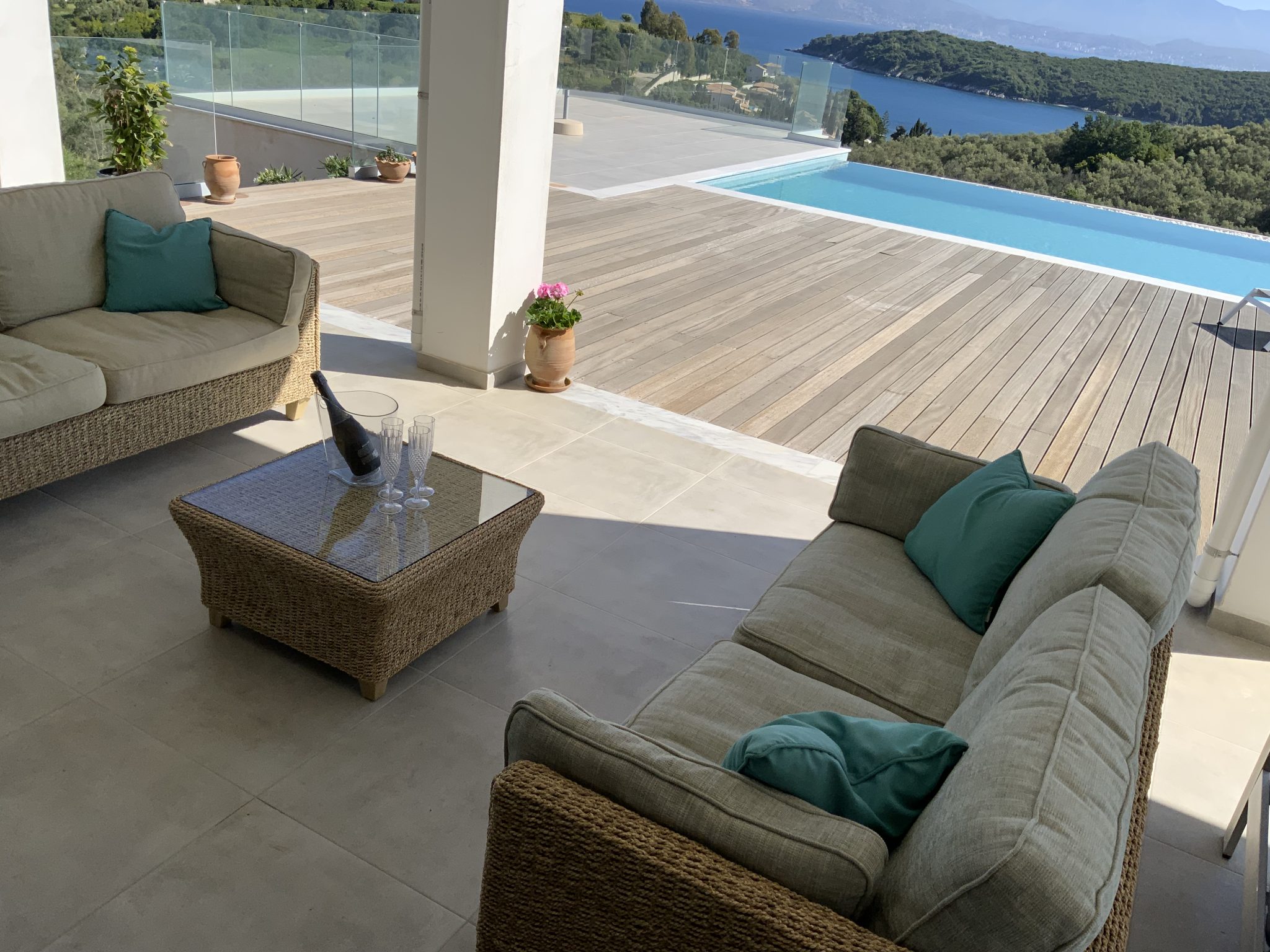 A modern outdoor patio with wicker furniture, turquoise cushions, and a glass-top table overlooks an infinity pool and a scenic view of green hills and a distant body of water, perfect for those seeking luxurious villa rentals in Corfu.