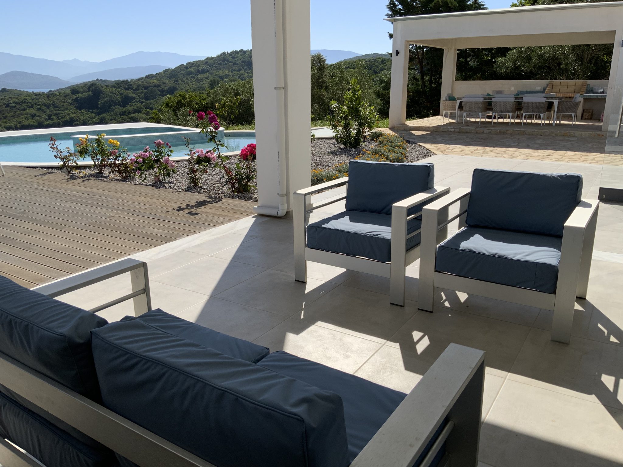 Outdoor seating area with blue cushions near a pool, flower bed, and distant mountains. A canopy covers a dining area with chairs and tables in the background. Bright, sunny day—perfect for those exploring holiday villas to rent in Corfu.