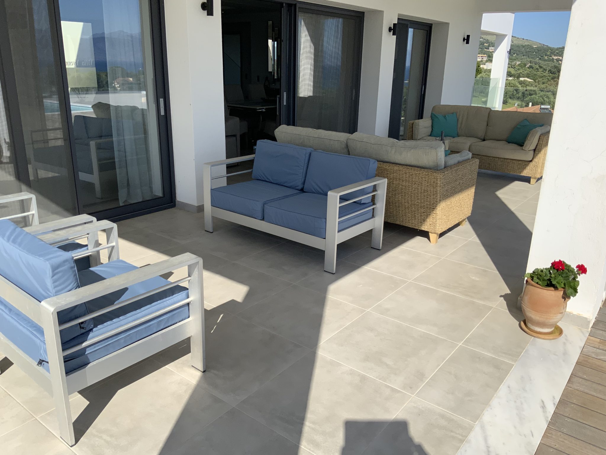 A modern outdoor patio with white framed furniture featuring blue seat cushions, a beige couch, and a wicker couch. A potted plant with red flowers adds charm in a corner, while mountains are visible in the distance, reminiscent of the scenic views from luxury Corfu villas.