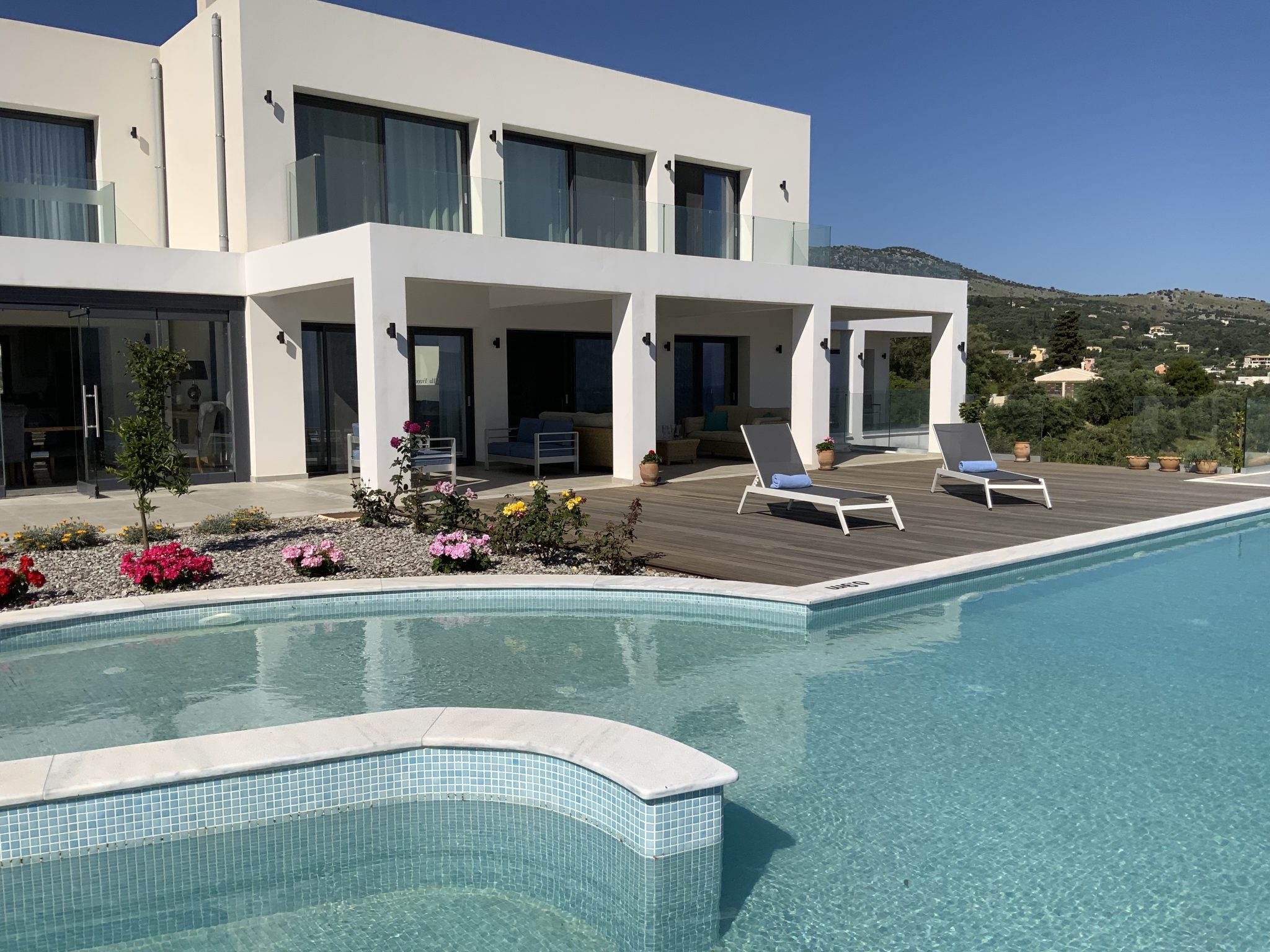 Modern white two-story house with large windows, patio, and pool area featuring wooden decking, two lounge chairs, and surrounding garden. This Corfu villa holiday spot boasts a green mountainous landscape under a clear blue sky in the background.