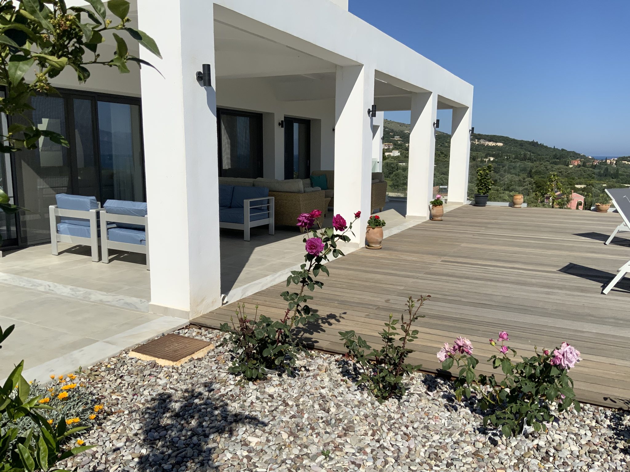 Modern white house with a spacious wooden deck, outdoor seating area, and a garden with blooming roses in the foreground. Clear blue sky in the background, reminiscent of idyllic Corfu villa holidays.