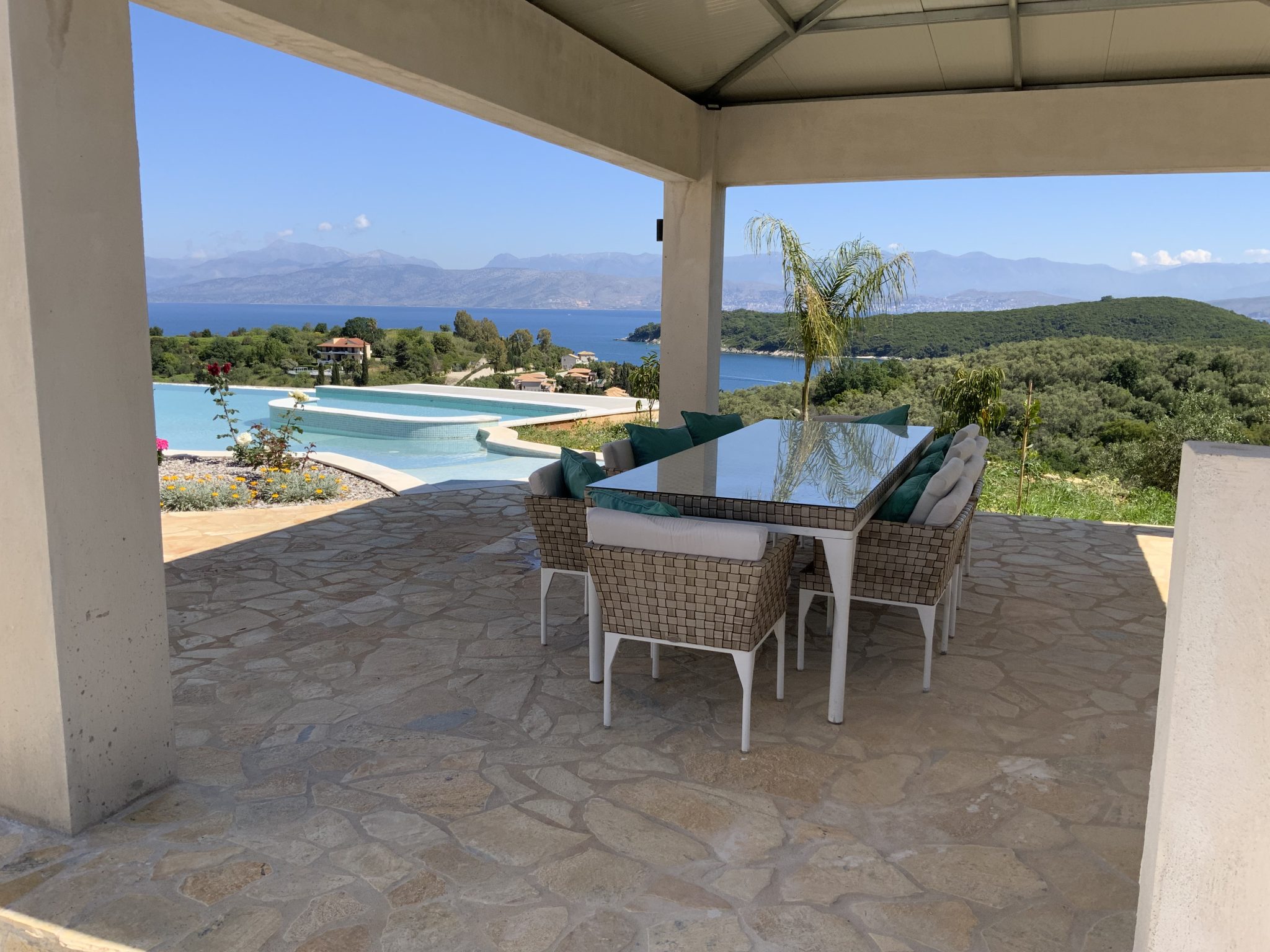 Covered patio with a glass dining table and wicker chairs, overlooking an infinity pool and scenic coastal landscape with mountains in the background, typical of villas in Corfu, Greece.