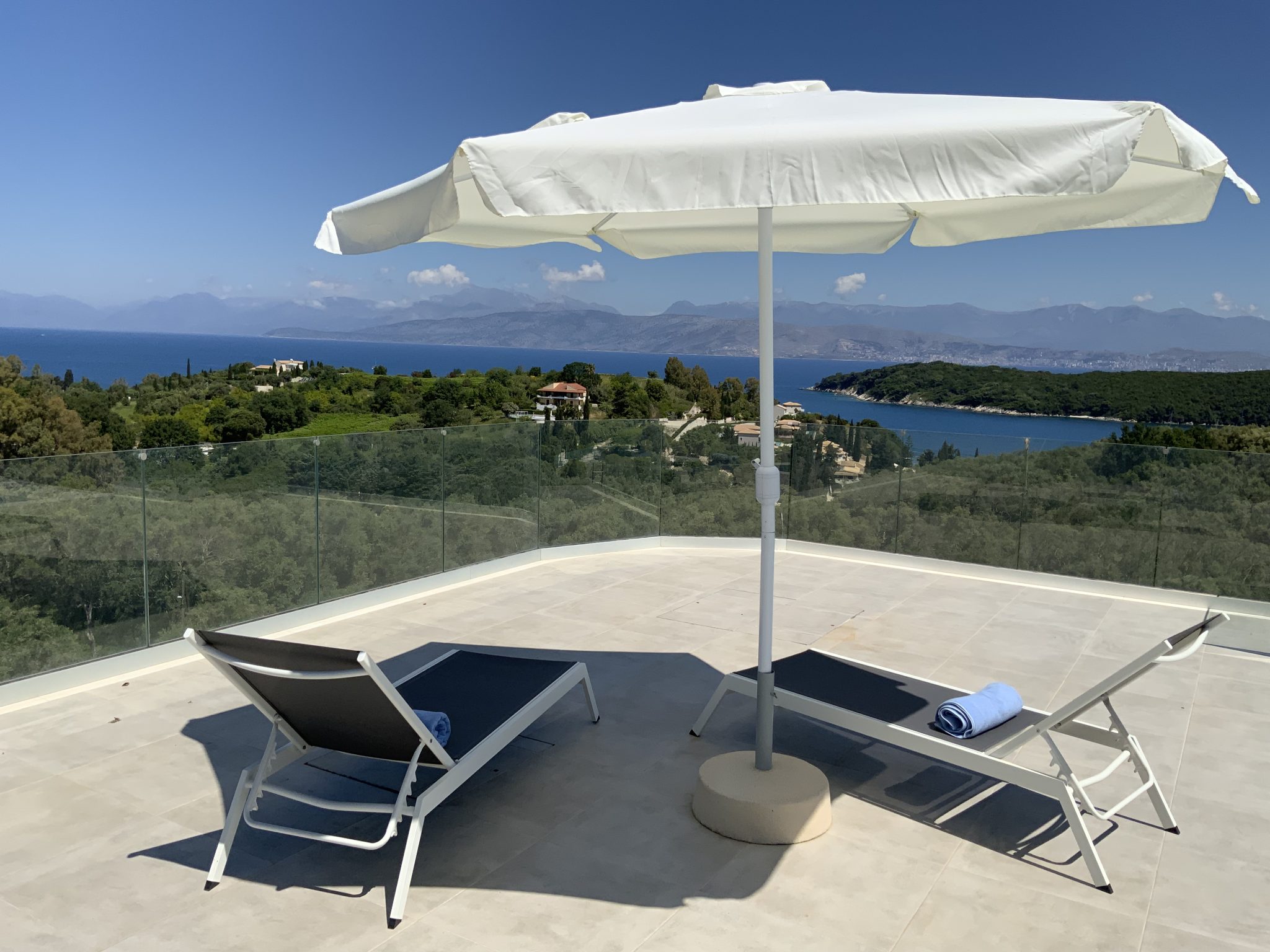 Two lounge chairs with towels sit under a large white umbrella on a patio, overlooking the scenic greenery, hills, and sea of Corfu, Greece under a clear blue sky.