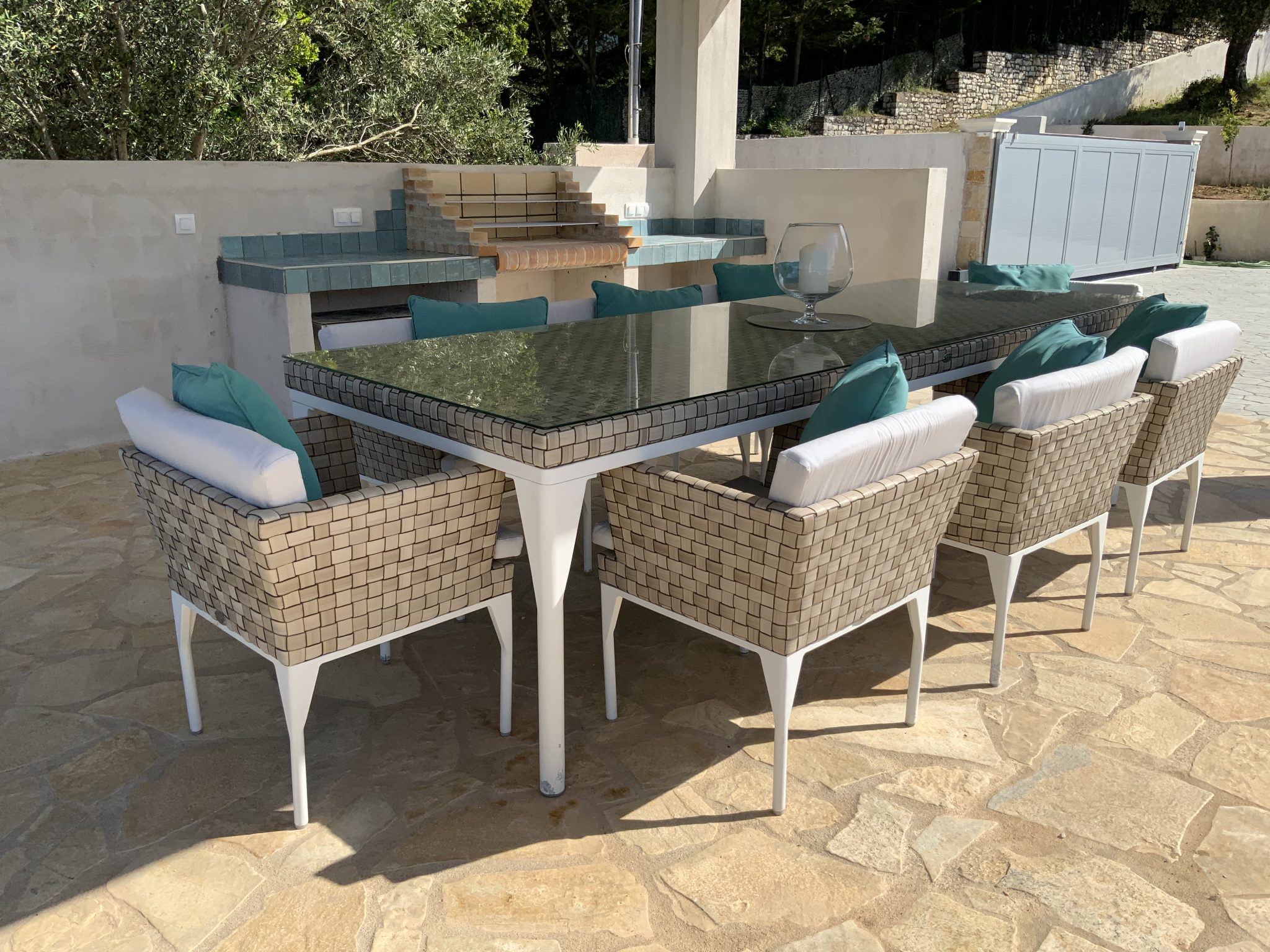 Outdoor patio area with a rectangular glass-top table and eight wicker chairs with white cushions and teal pillows, perfect for experiencing Corfu villa holidays. There is a built-in stone barbecue setup in the background.