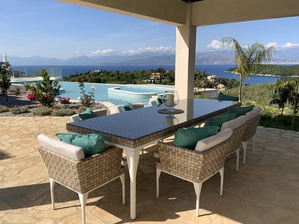 Outdoor dining area with wicker chairs and a glass-top table under a canopy, overlooking an infinity pool, lush greenery, the sea, and distant mountains—perfect for those seeking villas in Corfu with private pool access.