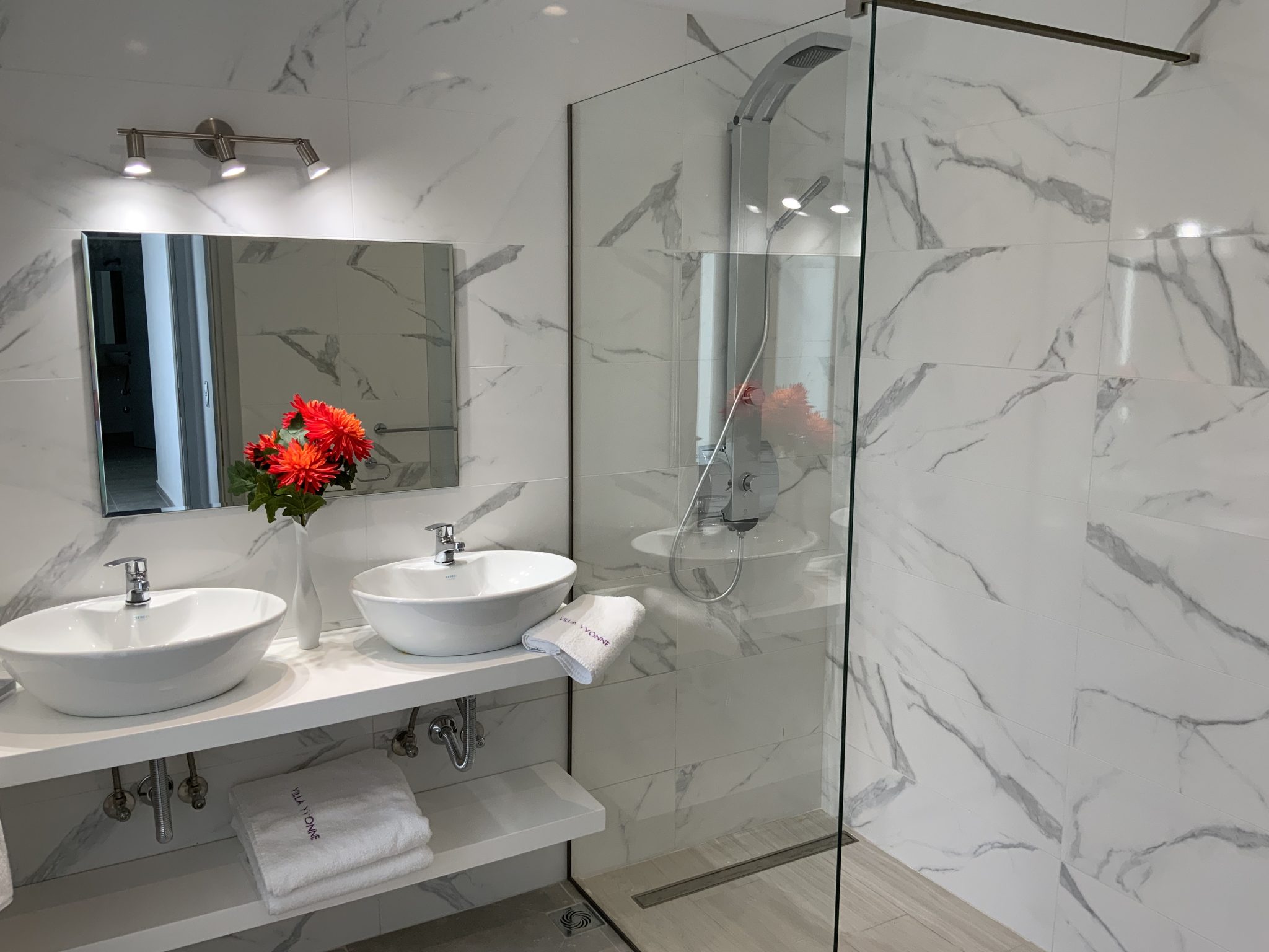 Modern bathroom with white marble walls, vanity with two sinks, mirrored cabinet, red flowers in a vase, and a glass-enclosed shower area in Villa Corfu. Towels are neatly arranged on the shelf below the sinks.