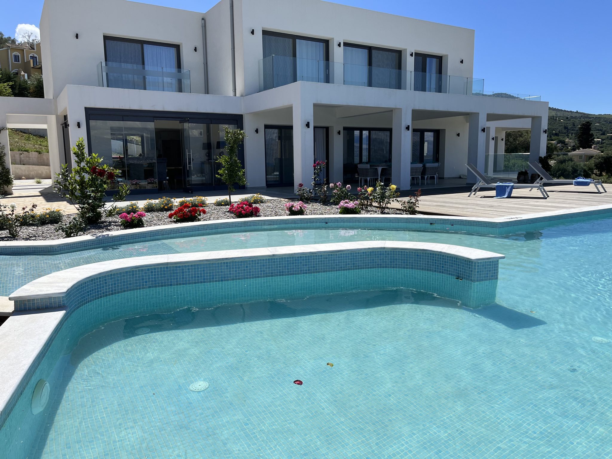 Modern two-story white house with large windows and balconies, featuring a landscaped garden and a blue tiled swimming pool in the foreground on a clear day. Perfect for those seeking luxurious villas to rent in Corfu.