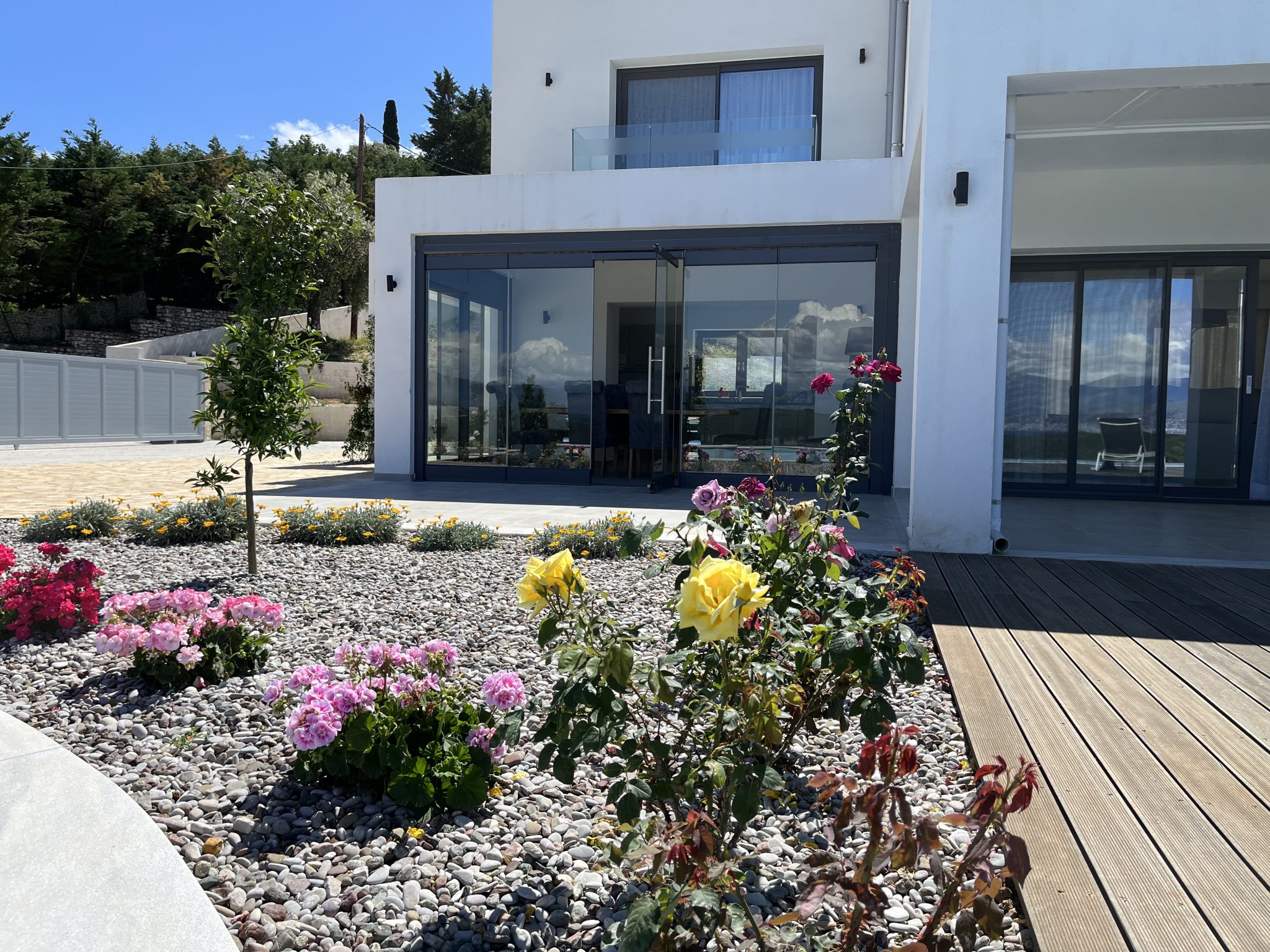 A modern white house with large glass doors opens to a patio. The foreground features a garden with blooming pink and yellow roses, set against a bed of gravel, and a wooden deck to the right—reminiscent of some luxurious villas to rent in Corfu.